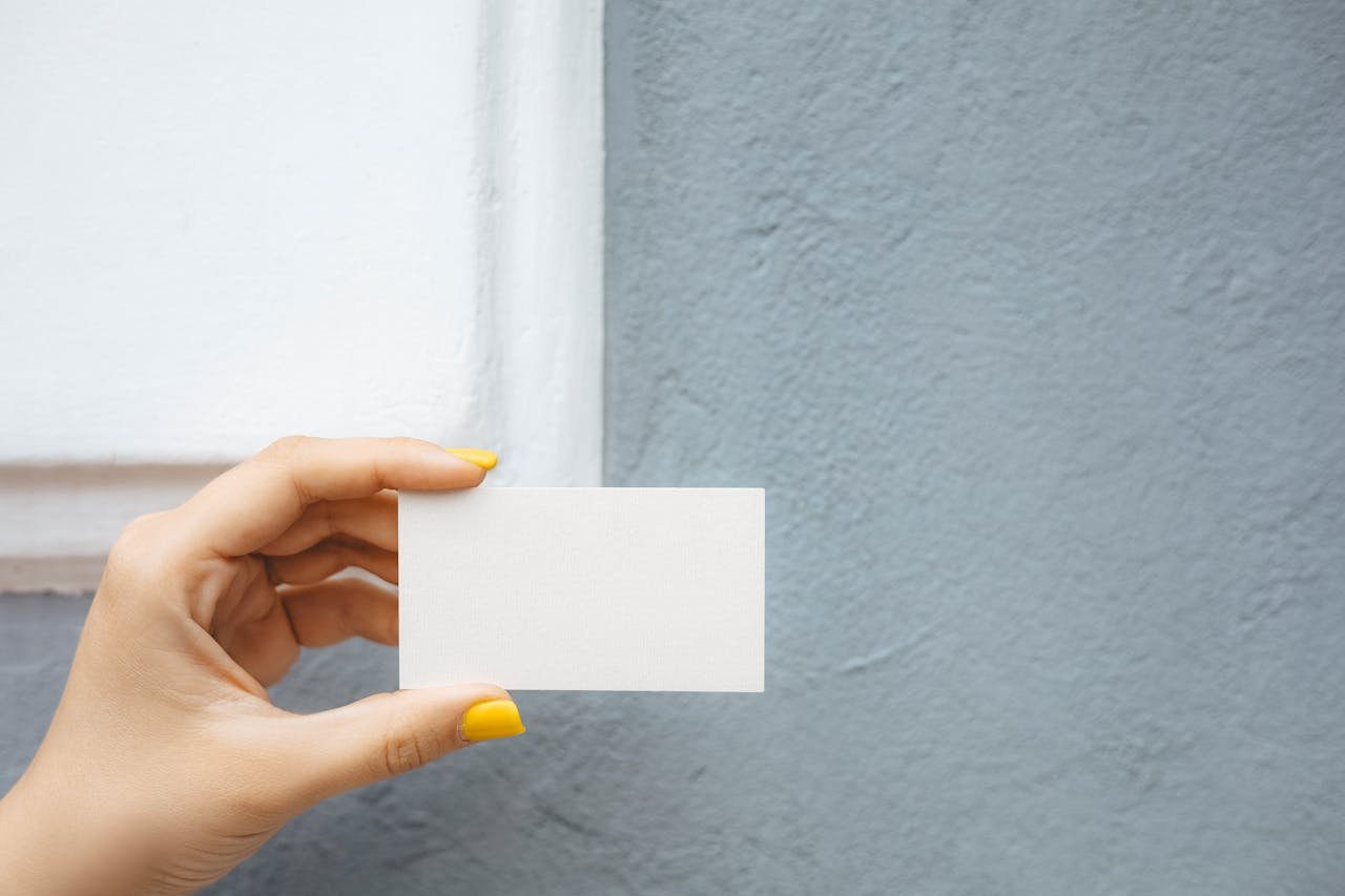 Close-up of a hand with yellow nail polish holding a blank white card against a textured blue wall.